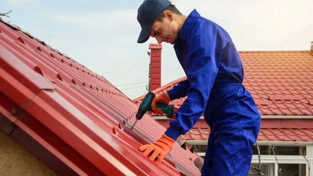 Roofer in blue uniform using a cordless drill to secure red metal roofing panels during routine maintenance.